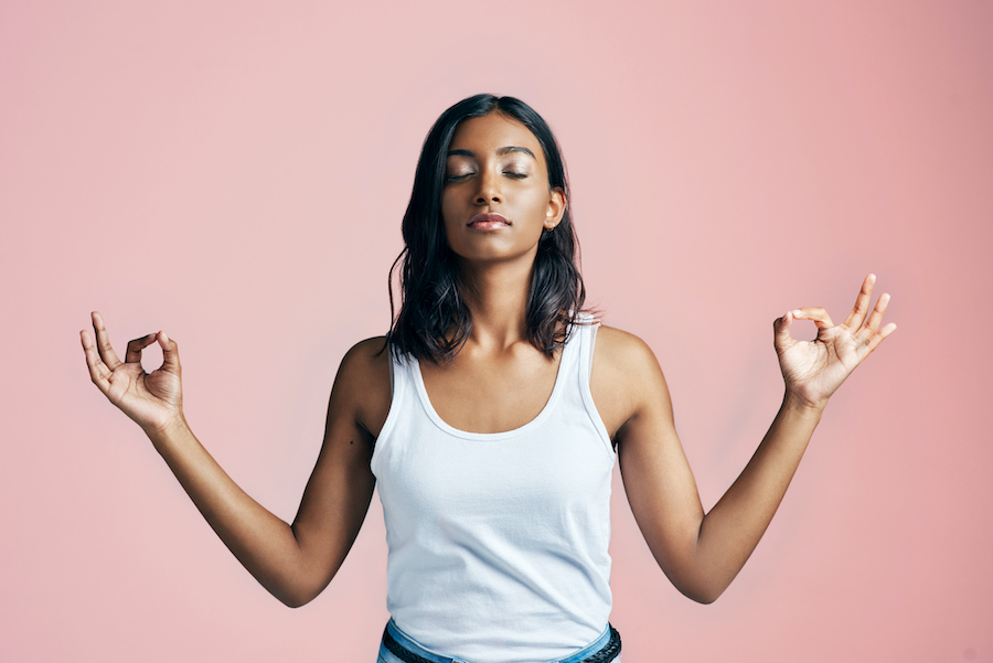 Brown woman in a white tank top stands in a zen pose trying to relax against a pink wall