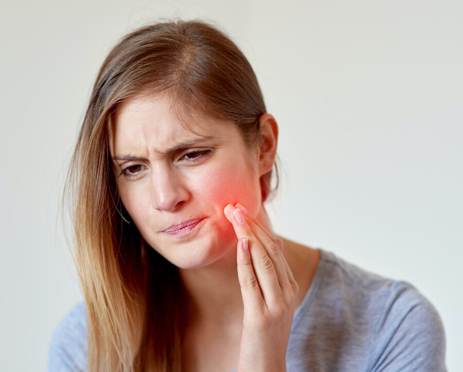 Blonde young woman cringes in pain and touches her cheek that is highlighted in red because of dry socket after oral surgery